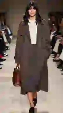 Modella in passerella con un trench marrone sopra un maglione color crema e una gonna grigia. Opzione vestiti per il cambio vestiti AI.