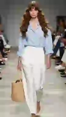 Modella in passerella con camicetta azzurra e pantaloni bianchi a gamba larga, con una borsa in tessuto. Opzione vestiti per il cambio vestiti AI.