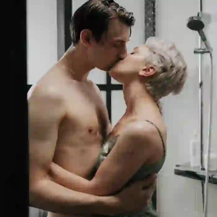 Foto eines küssenden Paares im Badezimmer. Der Mann trägt kein Hemd und die Frau trägt ein ärmelloses grünes Oberteil.