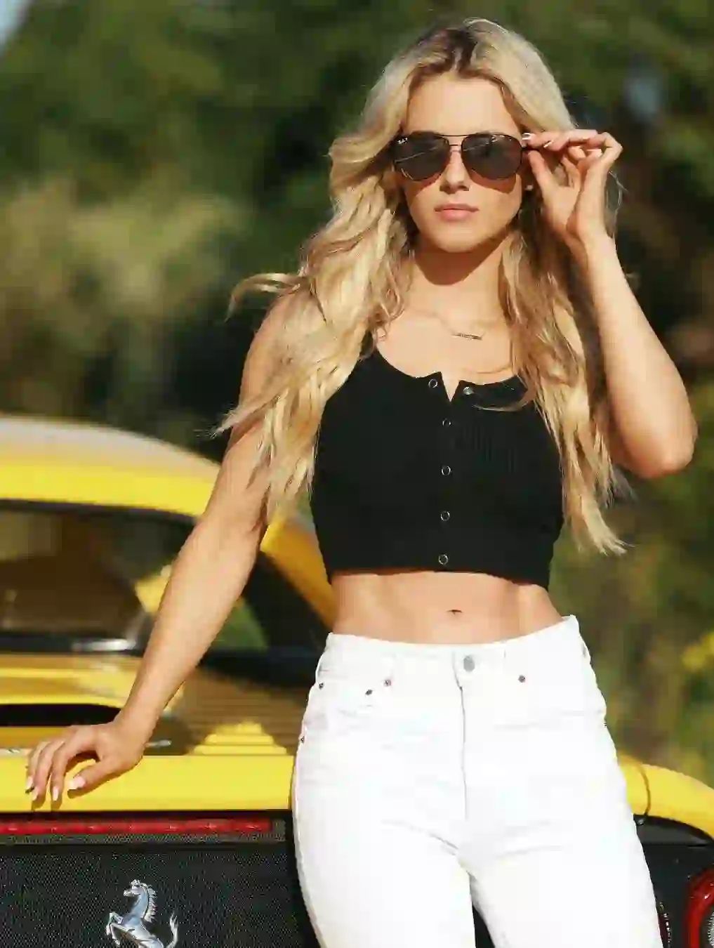 Photo of a woman with blonde wavy hair wearing sunglasses, a black tank top, and white pants, is standing against a yellow car.